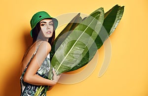 Beautiful young fashion girl with perfect skin in green hat hold tropical banana leaf in hands and covers