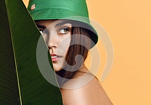 Beautiful young fashion girl with perfect skin in green hat hold tropical banana leaf in hands and covers