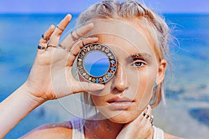 Beautiful young fashion model on the beach. Close up portrait of boho model holding small mirror at her eye