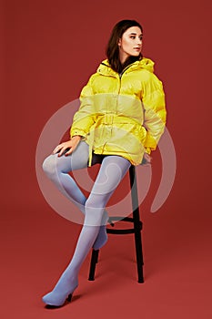 Beautiful young fashion woman in yellow oversized down jacket posing in studio on red background, using bar stool
