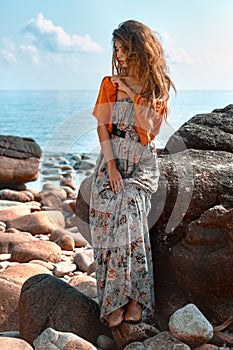 Beautiful young fashionable woman in elegant dress posing at the stone beach at sunset