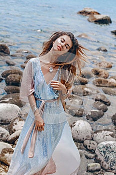 Beautiful young fashionable woman in elegant dress posing at the stone beach at sunset