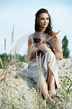 Beautiful young fashionable woman in elegant dress sitting on sand at sunset