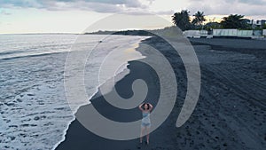 Beautiful young female on the beach of the ocean with black sand and waves at sunset on Bali, aircraft video 4K