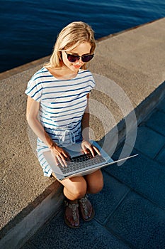 Beautiful young female blonde sitting on beach with laptop smiling and communicates over the Internet via Wi Fi