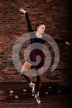 Beautiful young female classical ballet dancer on pointe shoes wearing a black leotard and skirt on a brick background