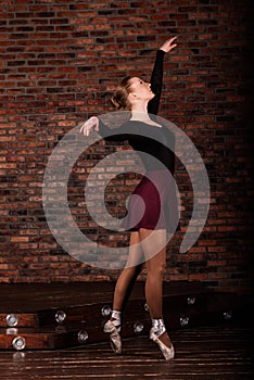 Beautiful young female classical ballet dancer on pointe shoes wearing a black leotard and skirt on a brick background