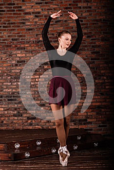 Beautiful young female classical ballet dancer on pointe shoes wearing a black leotard and skirt on a brick background