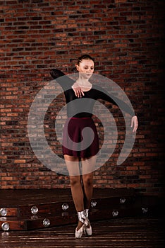 Beautiful young female classical ballet dancer on pointe shoes wearing a black leotard and skirt on a brick background