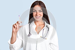 Beautiful young female doctor in white robe, has phonendoscope on neck, holds syringe with liquid, isolated over blue background,