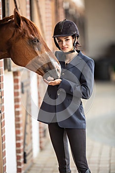 Beautiful young female jockey stands dressed up for the race next to its stabled paint horse