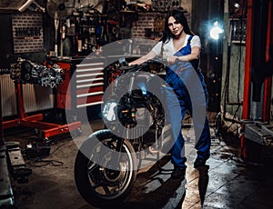 Beautiful young female mechanic in blue overalls posing with custom bobber in garage or workshop