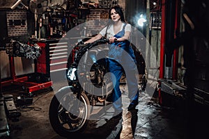 Beautiful young female mechanic in blue overalls posing with custom bobber in garage or workshop