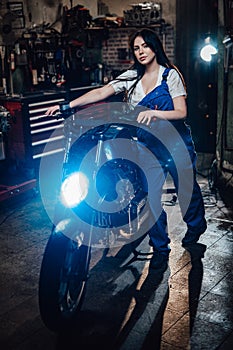 Beautiful young female mechanic in blue overalls posing with custom bobber in garage or workshop