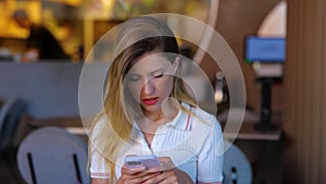 Beautiful young female sitting at table in restaurant and texting on mobile phone looking at camera. Close up of