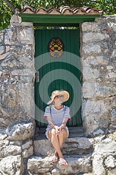 Beautiful young female tourist woman sitting and resting on vinatage wooden doorstep and textured stone wall at old