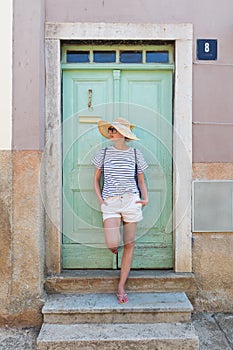 Beautiful young female tourist woman wearing sun hat, standing and relaxing in shade in front of vinatage wooden door in