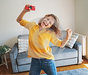 Beautiful young female in a yellow shirt is dancing happy and joyful in the living room with a smartphone in her hands, emotion