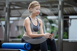 beautiful young fitness woman in sportswear sitting on staircase with yoga mat using smartphone While waiting training class