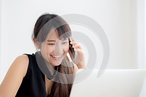 Beautiful young freelance asian woman smiling working on laptop computer and talking mobile smart phone at desk office