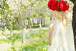 Beautiful young gentle elegant young blond woman with red peony in a wreath of white blouse walking in the lush apple orchard