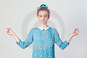 Beautiful young ginger girl doing yoga indoors at grey wall, keeping eyes closed, holding fingers in mudra gesture.