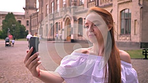Beautiful young ginger woman with bare shoulders having video chat and standing on street with building in background