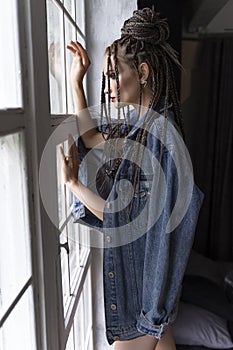 A beautiful young girl with an afro cornrows hairstyle, wearing a casual denim jacket and shorts, relaxes by the window and looks