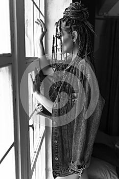 A beautiful young girl with an afro cornrows hairstyle, wearing a casual denim jacket and shorts, relaxes by the window and looks