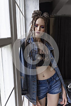 A beautiful young girl with an afro cornrows hairstyle, wearing a casual denim jacket and shorts, relaxes by the window and looks