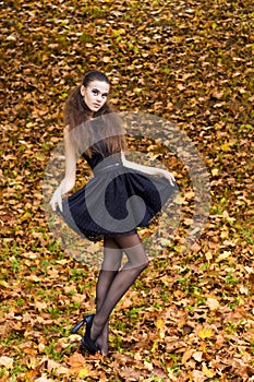 Beautiful young girl on the background of the leaves in autumn day on the street with fantasy makeup in a black dress