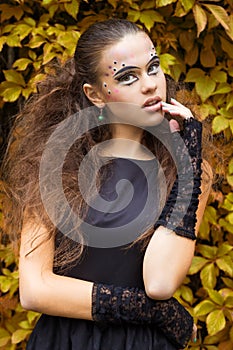 Beautiful young girl on the background of the leaves in autumn day on the street with fantasy makeup in a black dress