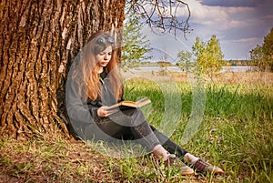 Beautiful young girl in dark clothes, sitting on the green grass near a large tree in the Park by the lake and reading a book unde