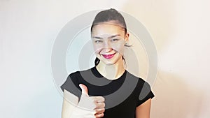 A beautiful young girl in a black t-shirt is smiling and showing thumb up looking at the camera.