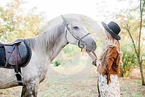 Beautiful young girl with blond hair in a suede jacket with a fringe , wearing black floppy hat, smiling and stroking her horse