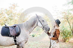 Beautiful young girl with blond hair in a suede jacket with a fringe , wearing black floppy hat, smiling and stroking her horse