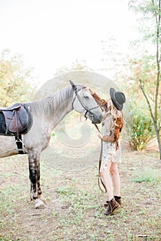 Beautiful young girl with blond hair in a suede jacket with a fringe , wearing black floppy hat, smiling and stroking her horse