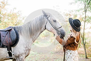 Beautiful young girl with blond hair in a suede jacket with a fringe , wearing black floppy hat, smiling and stroking her horse