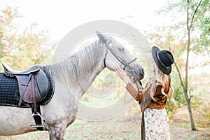 Beautiful young girl with blond hair in a suede jacket with a fringe , wearing black floppy hat, smiling and stroking her horse