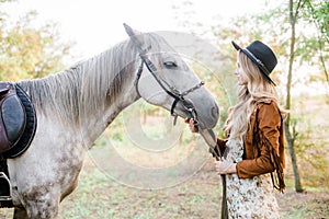 Beautiful young girl with blond hair in a suede jacket with a fringe , wearing black floppy hat, smiling and stroking her horse