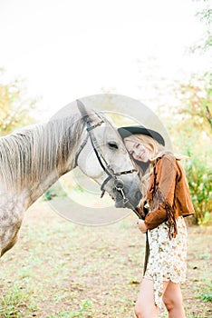 Beautiful young girl with blond hair in a suede jacket with a fringe , wearing black floppy hat, smiling and stroking her horse