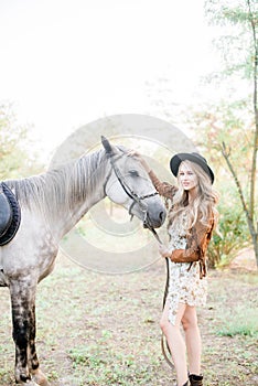 Beautiful young girl with blond hair in a suede jacket with a fringe , wearing black floppy hat, smiling and stroking her horse