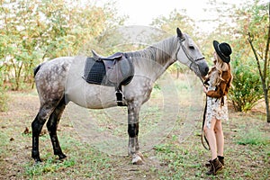 Beautiful young girl with blond hair in a suede jacket with a fringe , wearing black floppy hat, smiling and stroking her horse