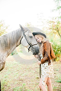 Beautiful young girl with blond hair in a suede jacket with a fringe , wearing black floppy hat, smiling and stroking her horse