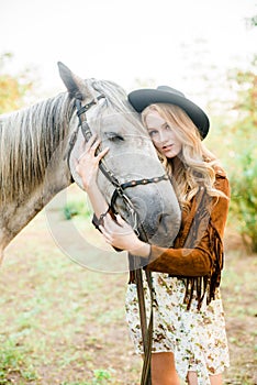 Beautiful young girl with blond hair in a suede jacket with a fringe , wearing black floppy hat, smiling and stroking her horse