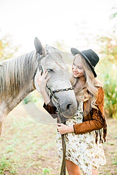 Beautiful young girl with blond hair in a suede jacket with a fringe , wearing black floppy hat, smiling and stroking her horse