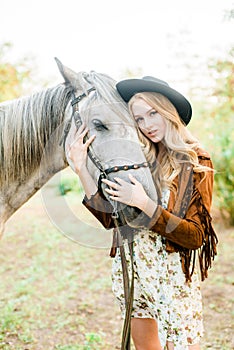 Beautiful young girl with blond hair in a suede jacket with a fringe , wearing black floppy hat, smiling and stroking her horse