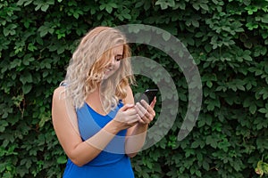 Beautiful young girl in a blue dress typing a message on the phone. Against the background of wild grapes, summer day. Copy space