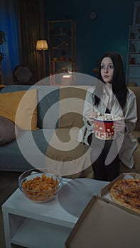 Beautiful young girl in casual clothing sitting on the sofa in the room watching movie eating popcorn. Vertical photo.