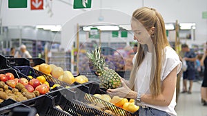 A beautiful young girl chooses a pineapple in the supermarket, sniffs it, checking for ripeness, and puts it in the cart